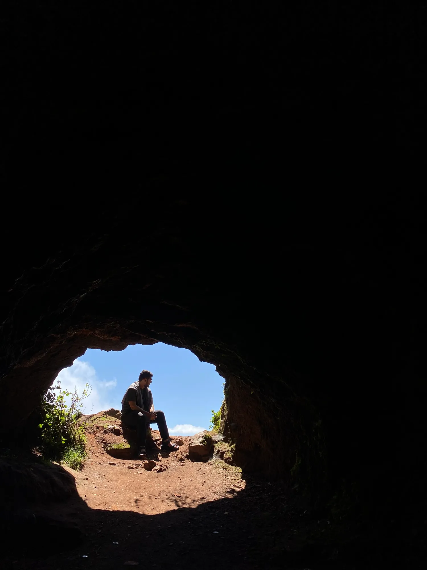 Suhruth silhouetted, sitting at the mouth of a dark cave against a bright blue sky.