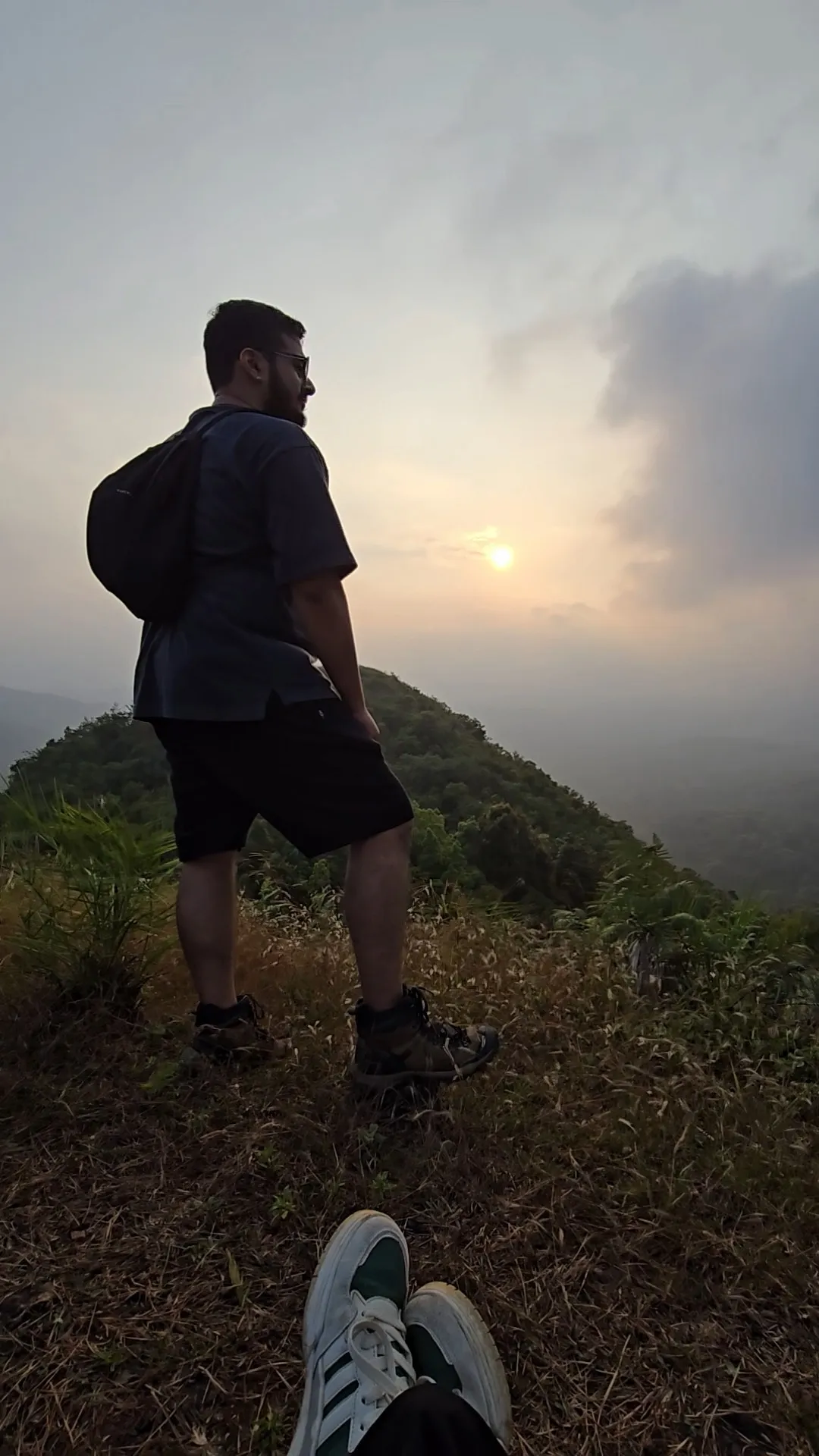 Suhruth with a backpack standing on a hilltop, looking out at a hazy sunset over forested hills.