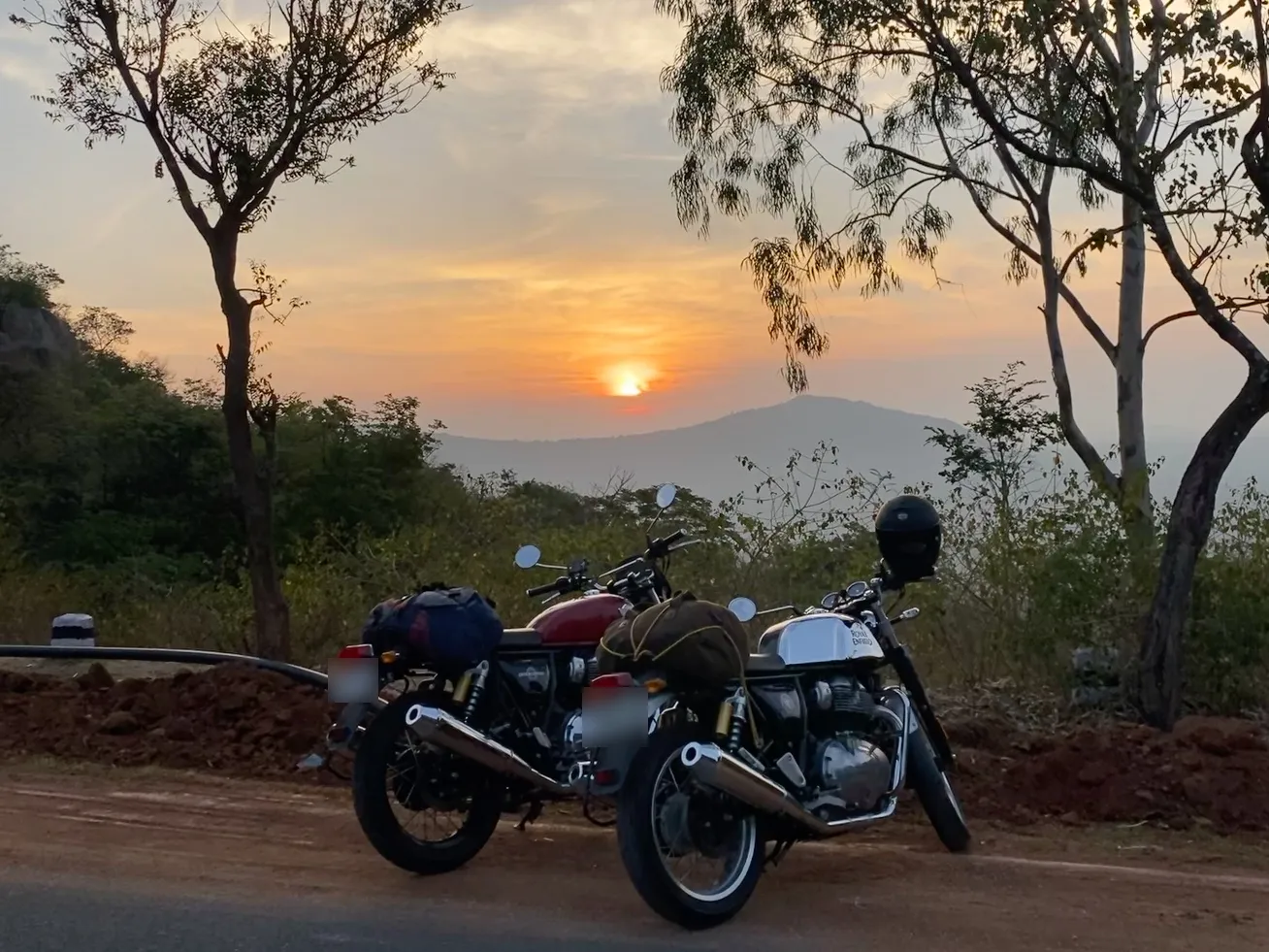 Two motorcycles, one red and one silver, parked at the edge of a hill road at sunset, with trees and a distant ridge behind them.