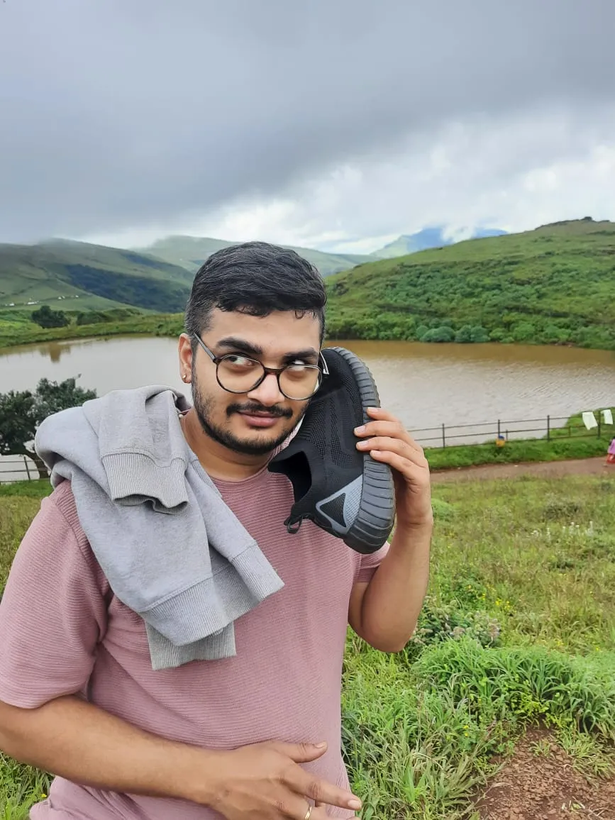 Suhruth by a lake in green hills, holding a black sneaker to his ear like a phone, a grey sweatshirt over his shoulder.