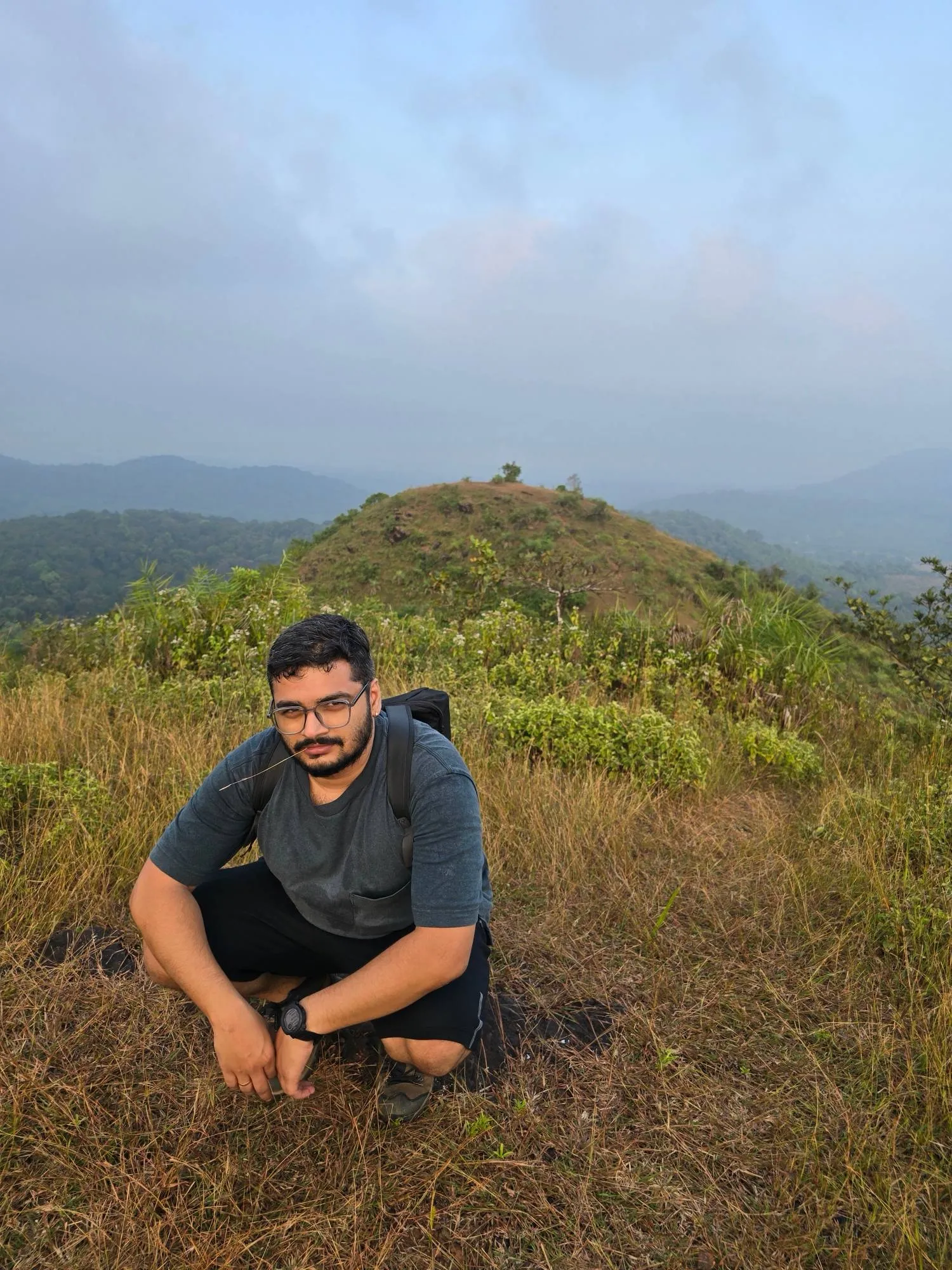 Suhruth crouching on a grassy hilltop with a backpack and a blade of grass in his mouth, forested hills and cloud behind him.